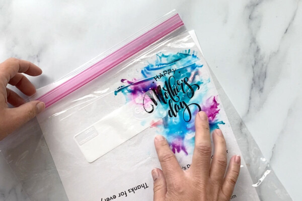 Bag with ink has been flipped over onto the paper, hand is blending the colors