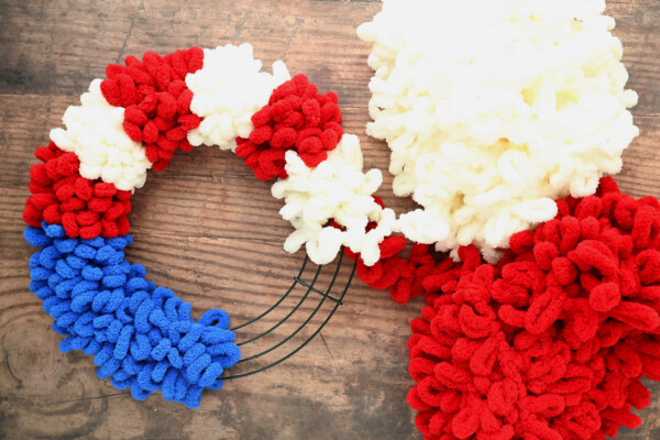 Wire wreath frame wrapped with blue loop yarn on one quarter; also wrapped with red and white loop yarn in stripes