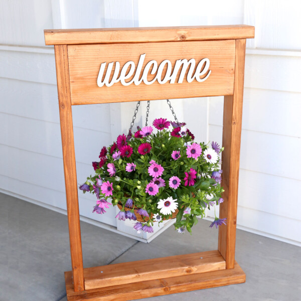 Wood plant stand with hanging flowers and the word welcome.