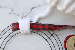 White yarn wrapped around wreath form three times, with ribbon under yarn.