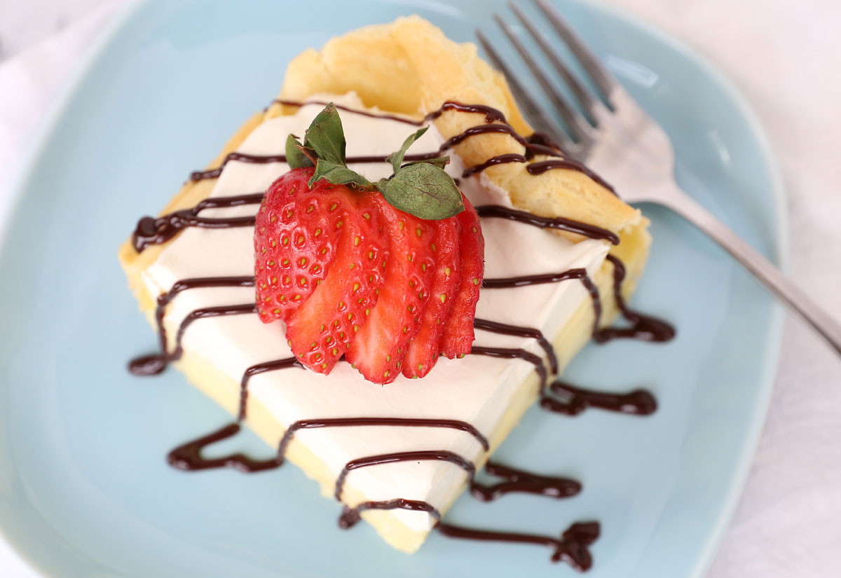Slice of cream puff cake on a plate with chocolate drizzle and strawberry.